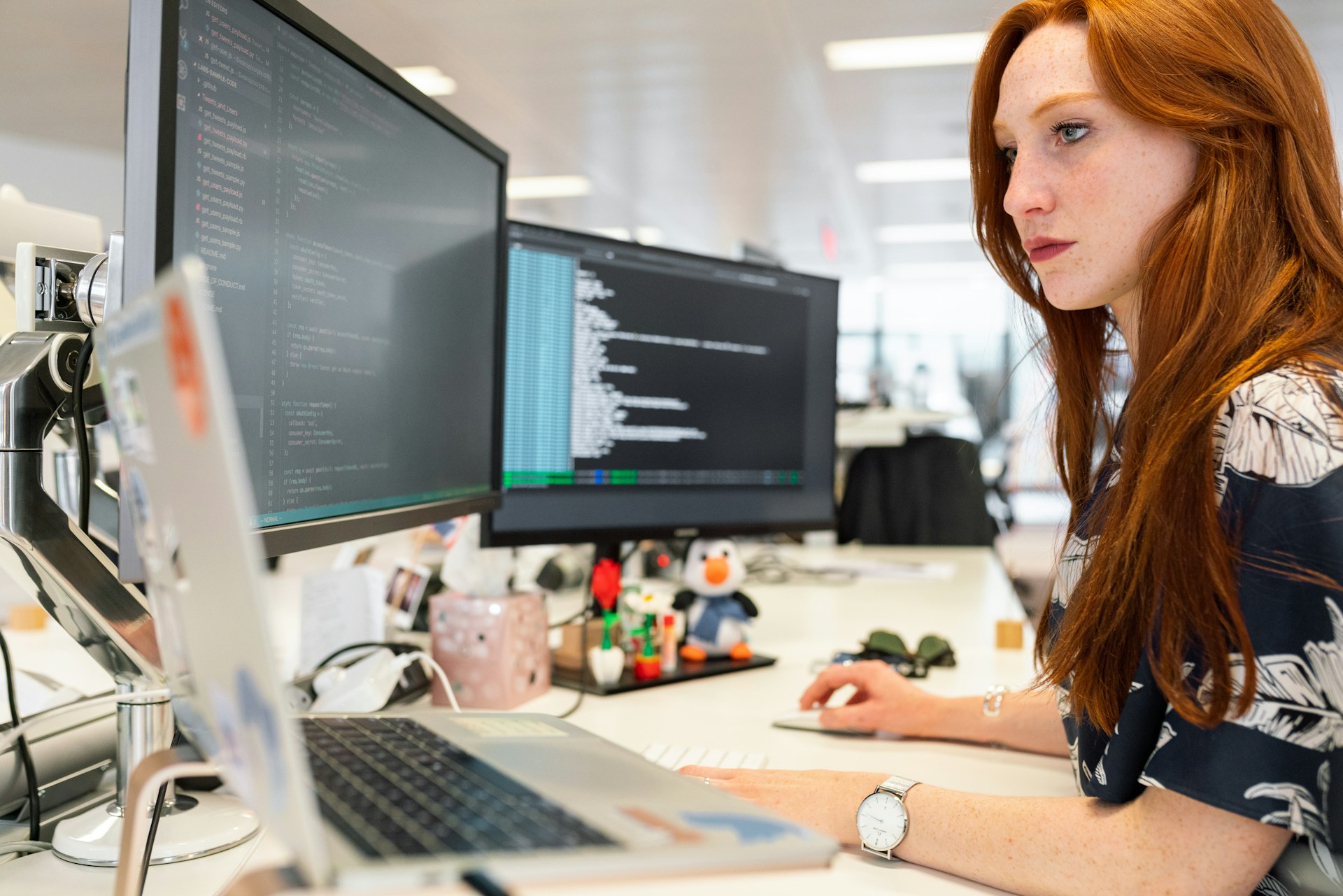 An image of a ginger haired lady sat in an office with 3 screens at their desk.